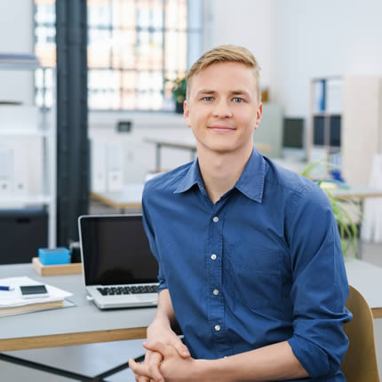 Junger Mann sitzt lächelnd an einem Bürotisch